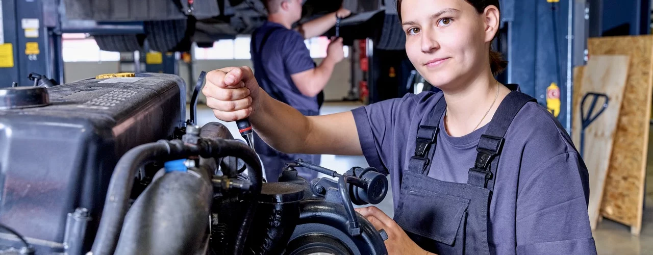 Person arbeitet mit einem Schraubendreher an einem Motor in einer Fahrzeugwerkstatt und blickt zur Kamera. Das Nutzfahrzeug befindet sich auf einer Hebeb&uuml;hne, w&auml;hrend im Hintergrund weitere Wartungsarbeiten stattfinden.