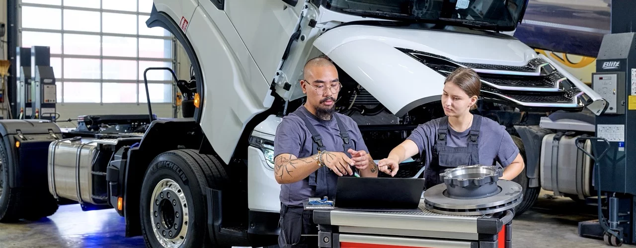 Zwei Personen besprechen technische Informationen an einem Laptop vor einem Nutzfahrzeug mit ge&ouml;ffneter Fahrerkabine. Auf einem Werkstattwagen liegt eine Bremsscheibe mit Radnabe.