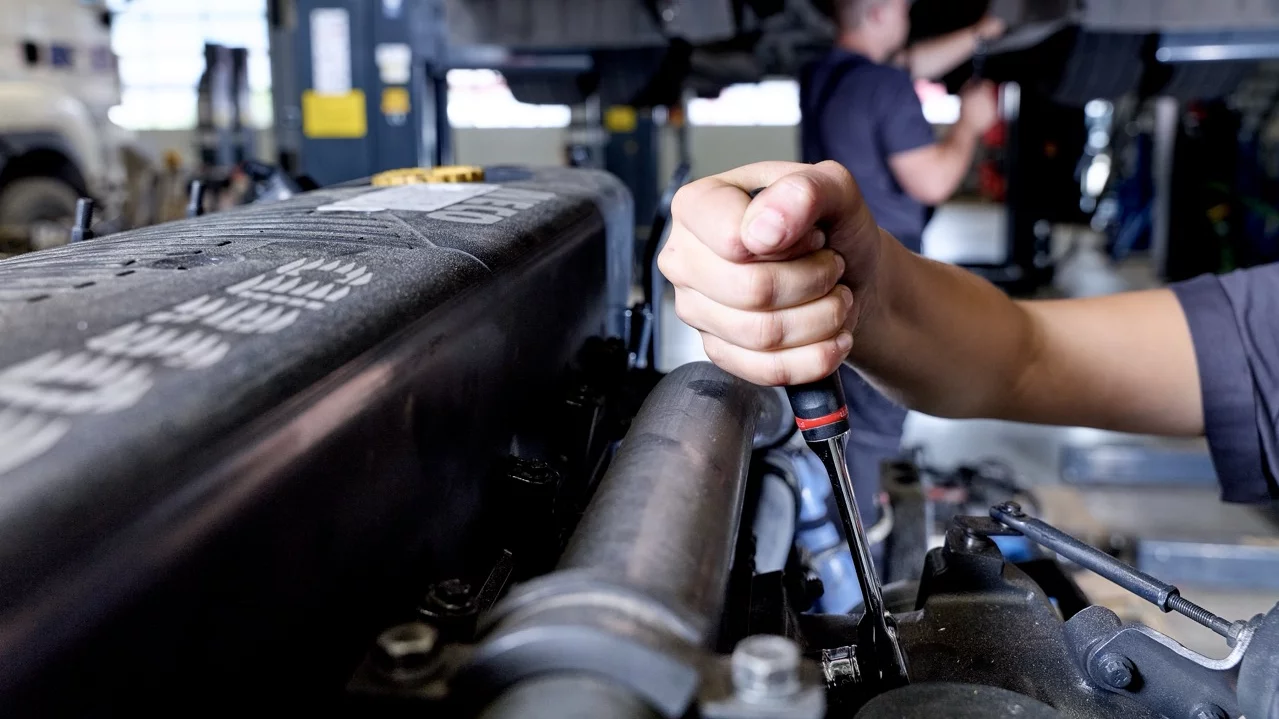 Nahaufnahme einer Hand mit Schraubendreher bei der Arbeit an einem Fahrzeugmotor. Im unscharfen Hintergrund sind ein angehobenes Nutzfahrzeug und eine weitere arbeitende Person in der Werkstatt zu sehen.