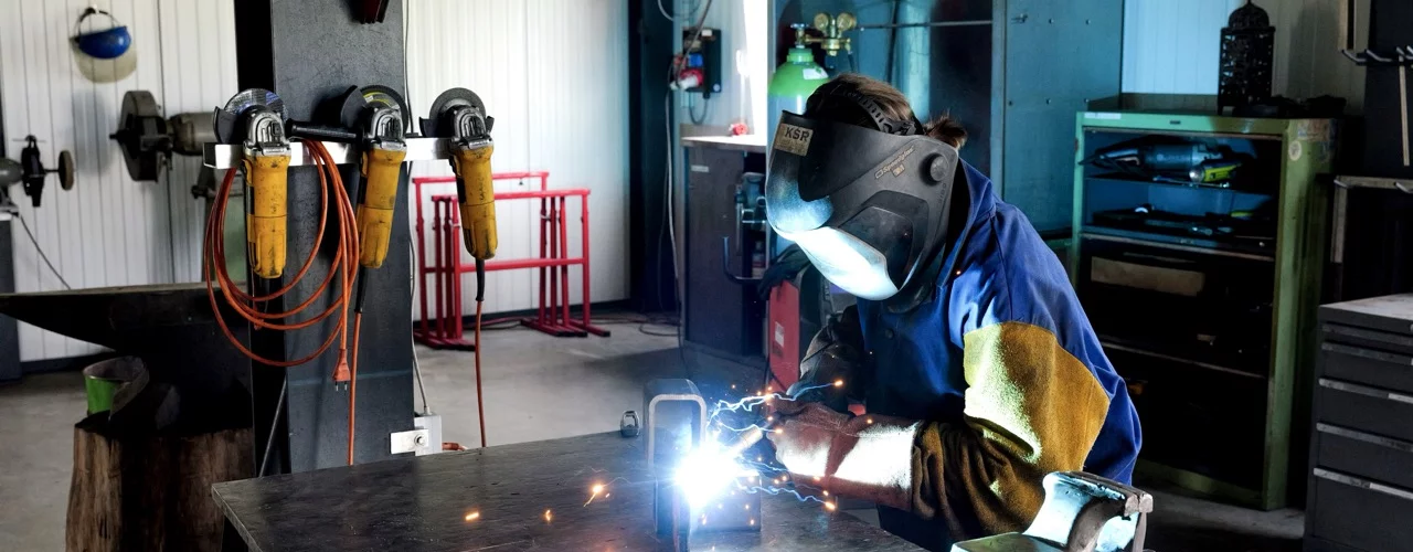 Person mit Schweisshelm und Schutzhandschuhen f&uuml;hrt Schweissarbeiten an einem Metallbauteil auf einer Werkbank durch. Funken entstehen am Schweisspunkt, w&auml;hrend Werkzeuge und Werkstatteinrichtungen im Hintergrund sichtbar sind.