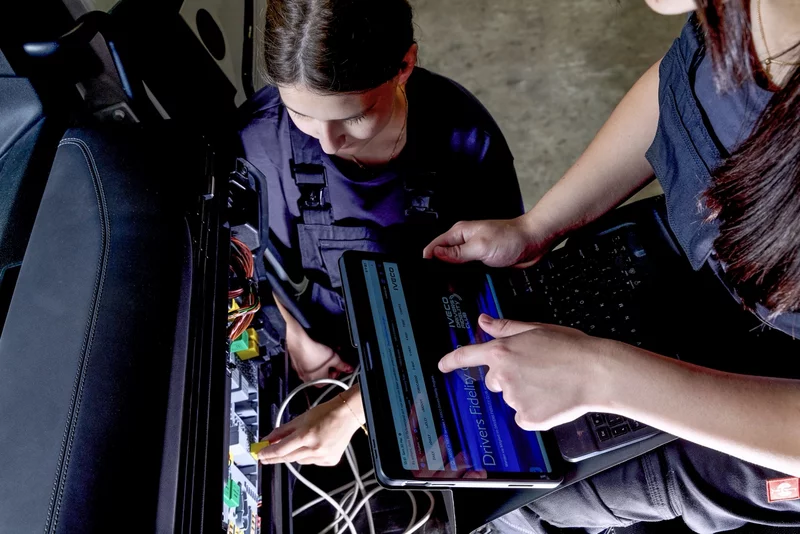 Zwei Personen arbeiten an der Fahrzeugelektronik. Eine Person pr&uuml;ft elektrische Verbindungen in einem ge&ouml;ffneten Technikfach, w&auml;hrend eine weitere Person Daten auf einem Tablet auswertet.