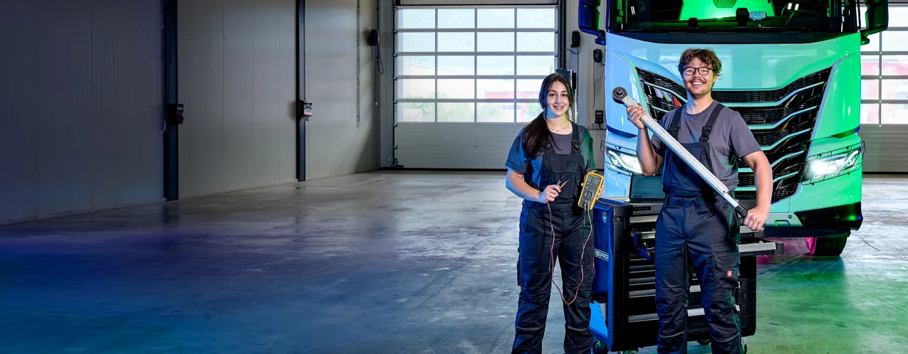Zwei Personen in Arbeitskleidung stehen mit Diagnose- und Servicewerkzeugen vor einem modernen Lastwagen in einer Nutzfahrzeugwerkstatt. Zwischen ihnen befindet sich ein mobiler Werkstattwagen mit Mess- und Diagnoseger&auml;ten.