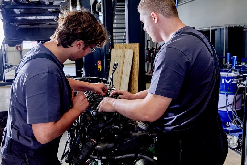 Zwei Personen arbeiten an einem ausgebauten Fahrzeugmotor in einer Werkstatt. Verschiedene Motorkomponenten und technische Einrichtungen sind im Arbeitsbereich sichtbar.
